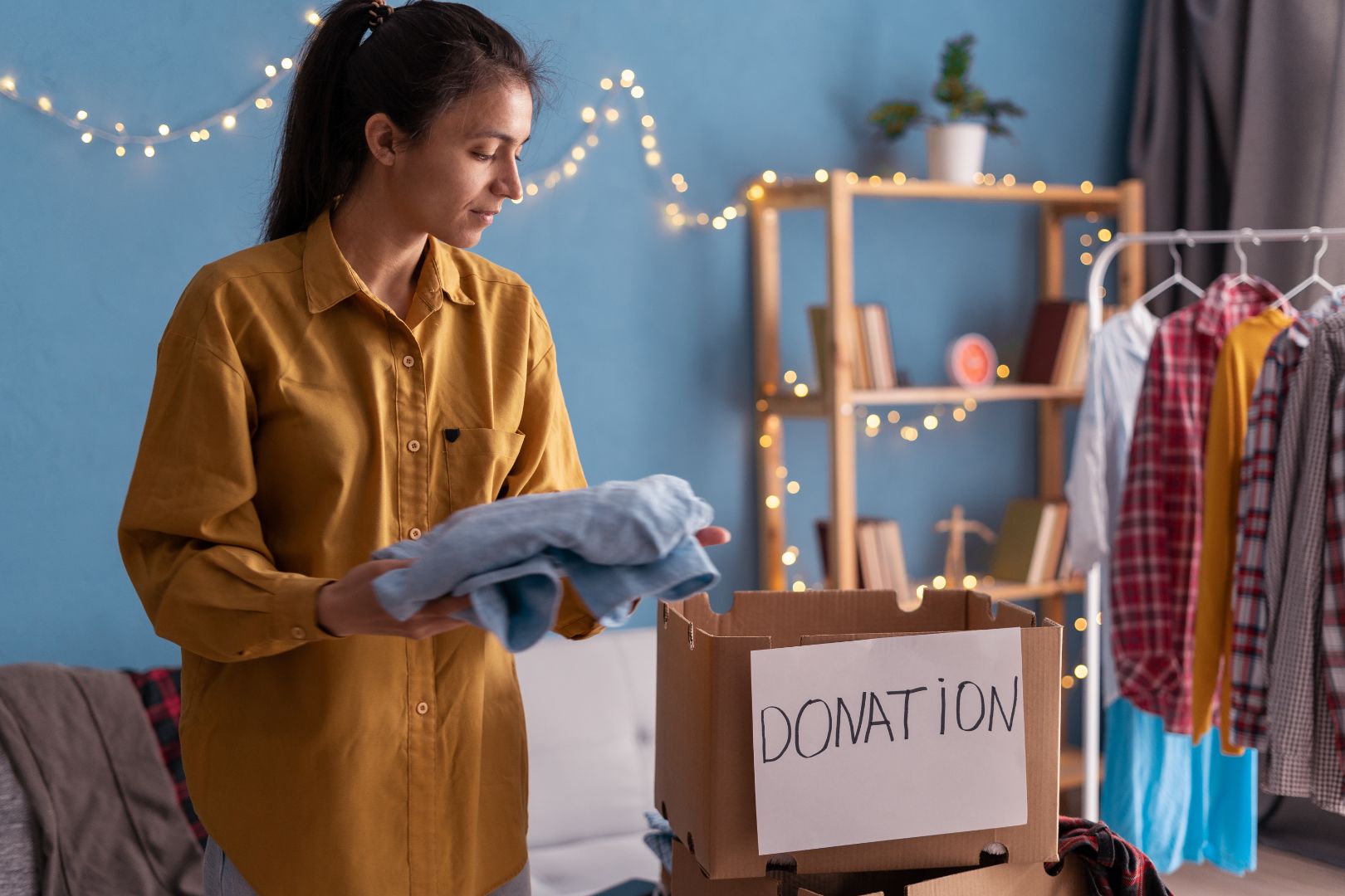 Woman is holding a folded blue shirt and she’s about to place it in a donation box. There is a rack of clothes near her.