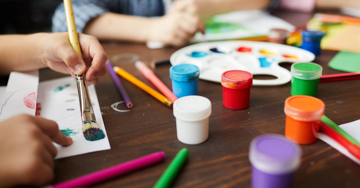 Close-up of children using art supplies at a table. Paint, canvases, and other coloring tools are near the children.