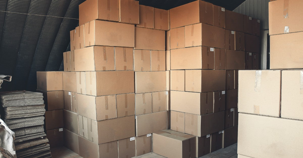 Cardboard boxes stacked on top of each other in a storage room. The boxes have clear tape and white stickers on them.