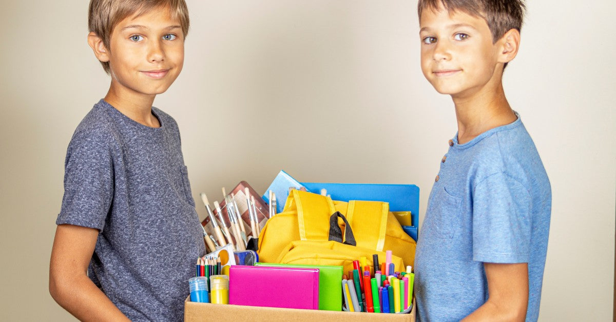 Two young boys holding a box of school supplies. The supplies include colored folders, markers, paintbrushes, and more.