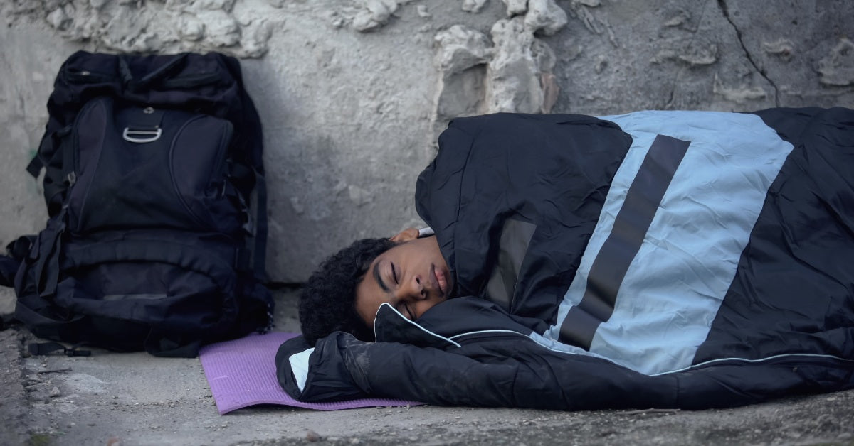 An African American man is sleeping outside in a black and blue sleeping bag. A black backpack is near his head.