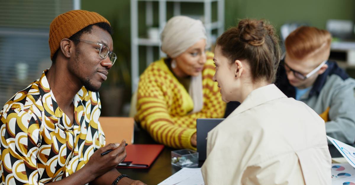 An African American man and a white woman collaborating on something. They're sitting at a table with other people.