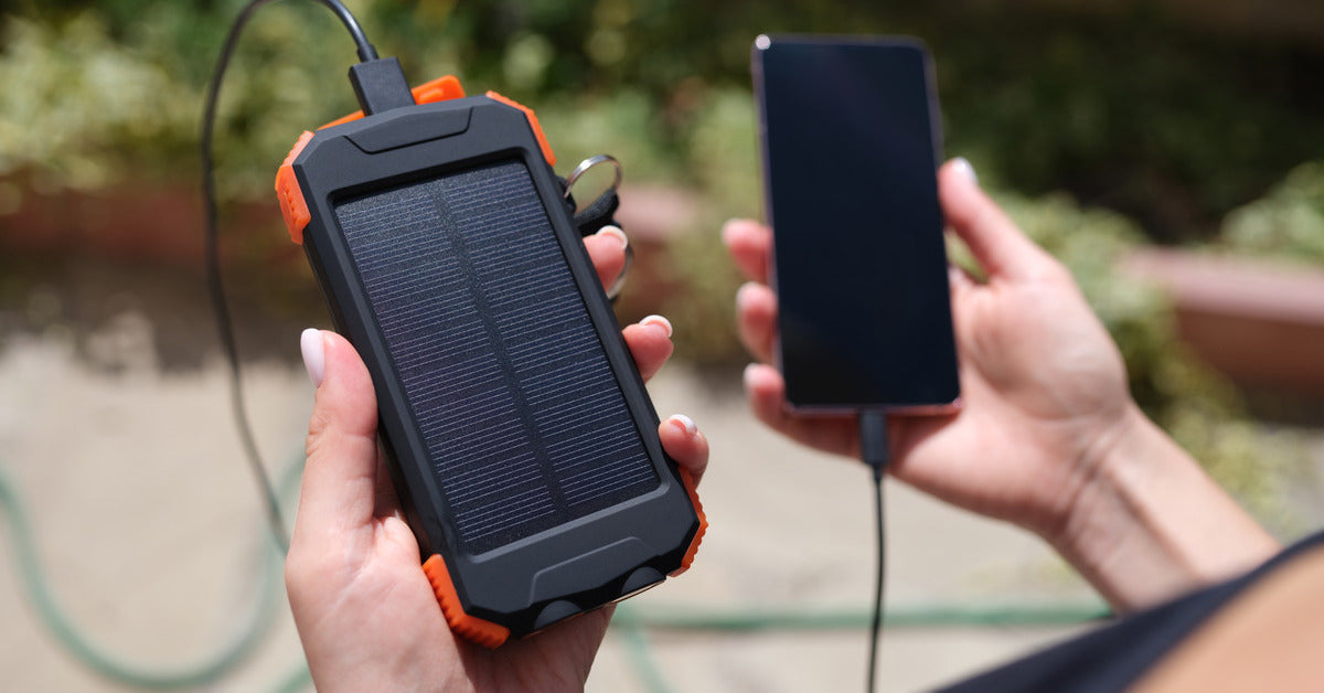 Someone holding a black and orange solar-powered phone charger and their black smartphone. A black cord connects the devices.