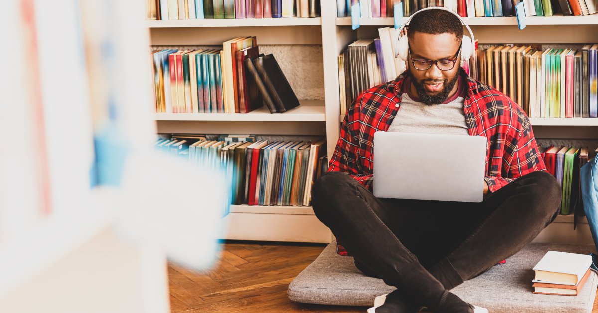 A Black man sits on the floor in front of a library bookshelf. He wears headphones and looks at his computer.